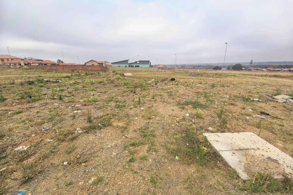 A vacant residential plot featuring natural ground cover, a concrete inspection cover, and surrounding perimeter walling in a developing neighborhood.