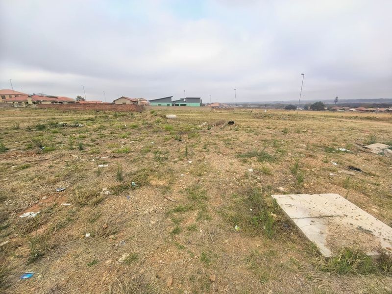 A vacant residential plot featuring natural ground cover, a concrete inspection cover, and surrounding perimeter walling in a developing neighborhood.