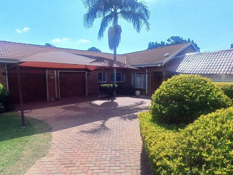 Front view of a single-story brick house featuring a paved driveway, double garage, and established garden with a shade sail.