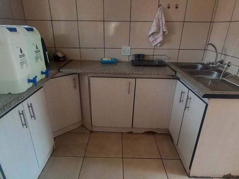 A functional kitchen area featuring tiled splashbacks, laminate countertops, and double stainless steel sink, with cabinetry showing signs of wear and water damage.