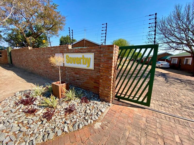 Entrance view of the Soverby townhouse complex featuring a brick boundary wall, electric fencing, and a secure metal gate leading to the paved driveway.