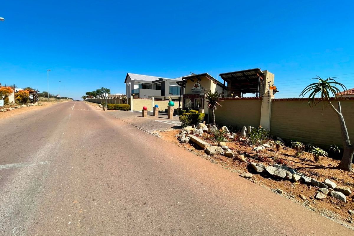 Street view of the residential estate entrance featuring a secure gatehouse, perimeter walling, and landscaped verge along the paved access road.