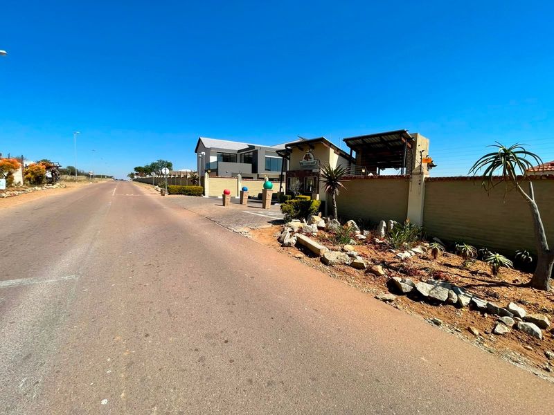 Street view of the residential estate entrance featuring a secure gatehouse, perimeter walling, and landscaped verge along the paved access road.