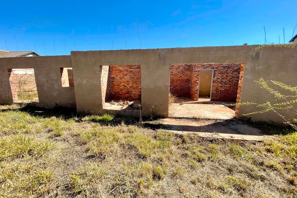 An unfinished residential structure showing exposed brick walls and concrete foundation on a vacant plot of land.