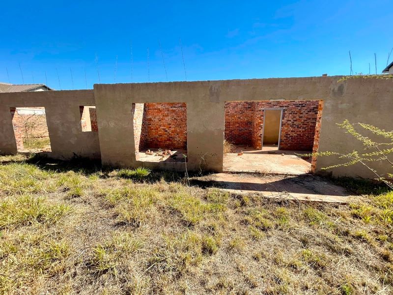 An unfinished residential structure showing exposed brick walls and concrete foundation on a vacant plot of land.