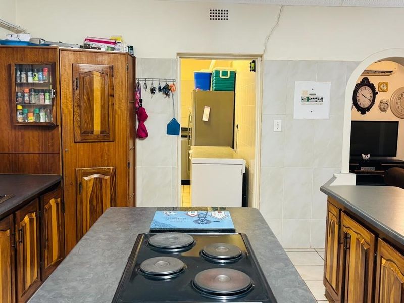 A dated kitchen featuring wooden cabinetry, laminate countertops, and a central island with an electric hob, leading through an archway to a scullery area.