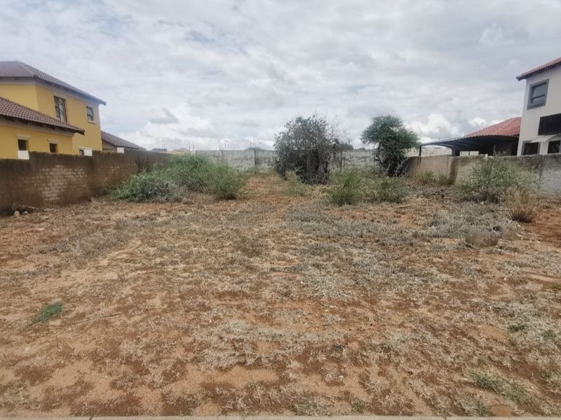 A vacant residential plot featuring perimeter boundary walls and natural ground cover, situated between existing residential properties.