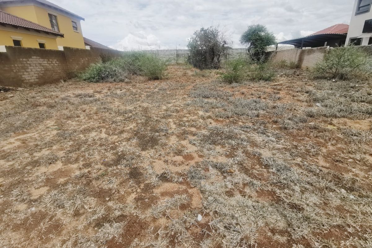 A vacant residential plot enclosed by perimeter boundary walls, featuring natural ground cover and scattered shrubs.