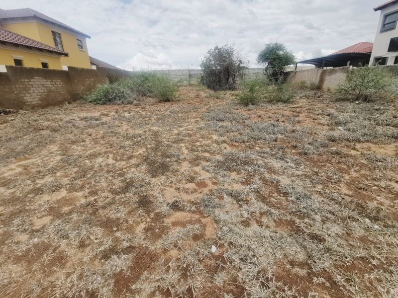 A vacant residential plot enclosed by perimeter boundary walls, featuring natural ground cover and scattered shrubs.