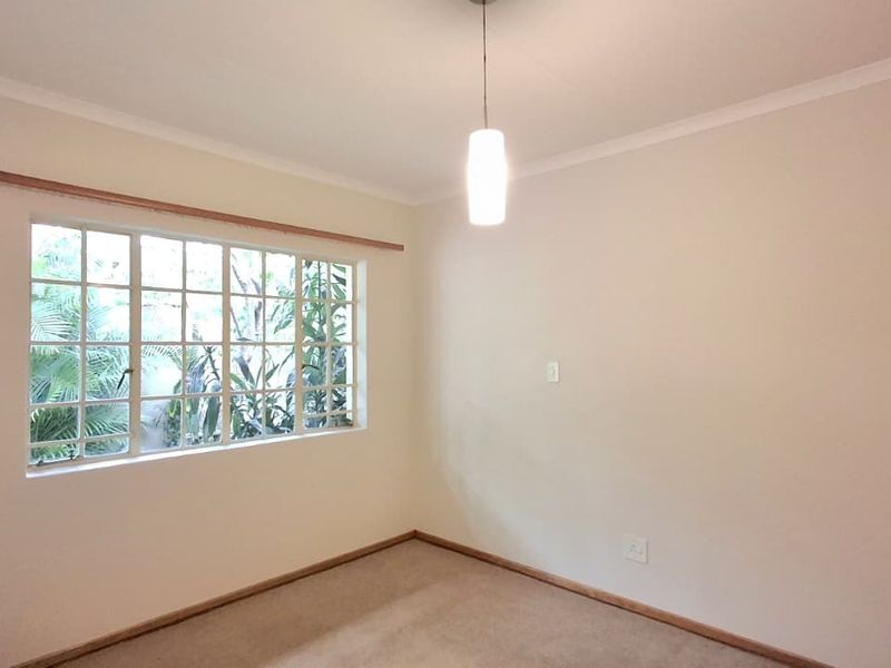 A bright, empty bedroom featuring carpeted flooring, a multi-pane window with a wooden curtain rod, and a central pendant light fixture.
