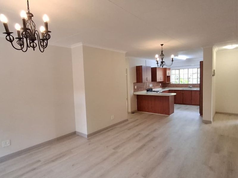 Spacious open-plan living and dining area flowing into a functional kitchen with wood-finish cabinetry, peninsula counter, and modern laminate flooring throughout.