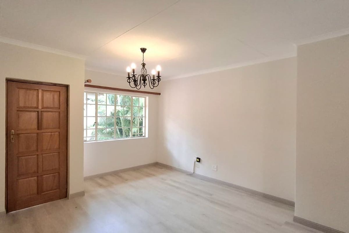 A bright, empty living room featuring light-toned laminate flooring, a classic timber panel door, and a decorative wrought iron chandelier.