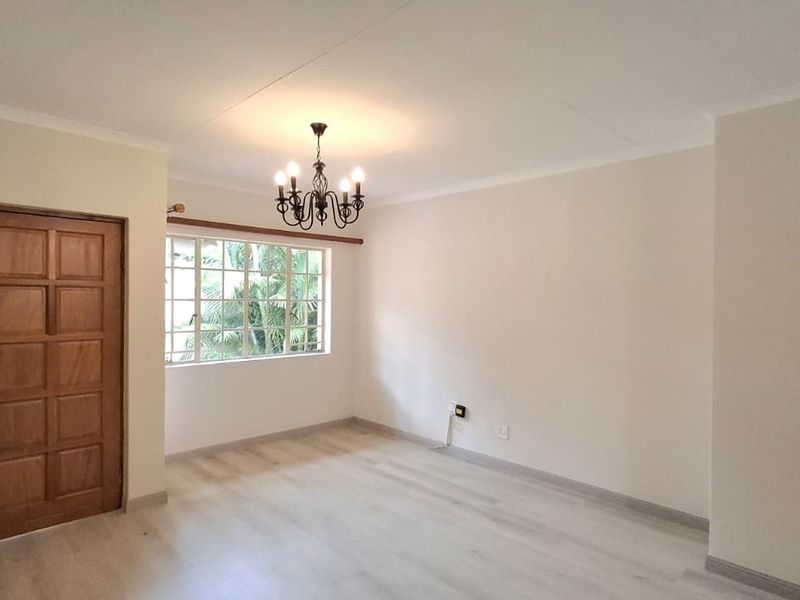 A bright, empty living room featuring light-toned laminate flooring, a classic timber panel door, and a decorative wrought iron chandelier.