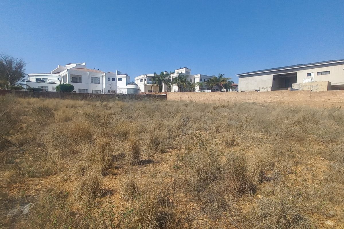 A view of a vacant residential plot featuring dry vegetation and a boundary wall, with neighboring residential properties visible in the background.