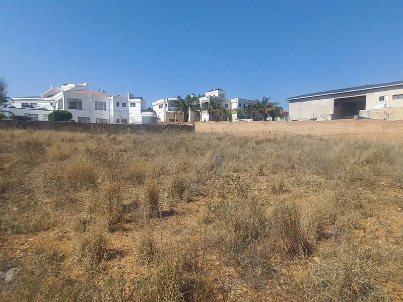 A view of a vacant residential plot featuring dry vegetation and a boundary wall, with neighboring residential properties visible in the background.