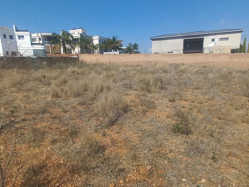A vacant residential plot featuring dry vegetation and a perimeter boundary wall, situated in an established neighborhood.