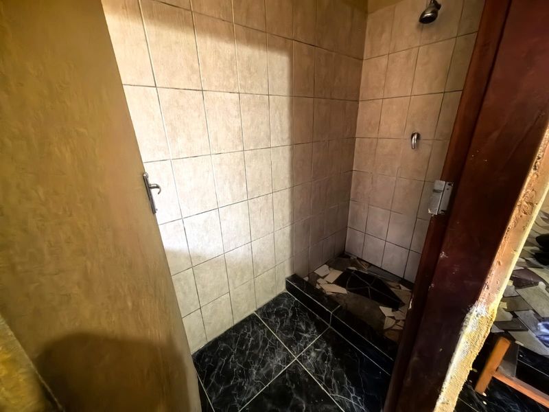 A small shower enclosure featuring tiled walls and flooring, currently showing signs of wear and damage to the floor tiles and door frame.