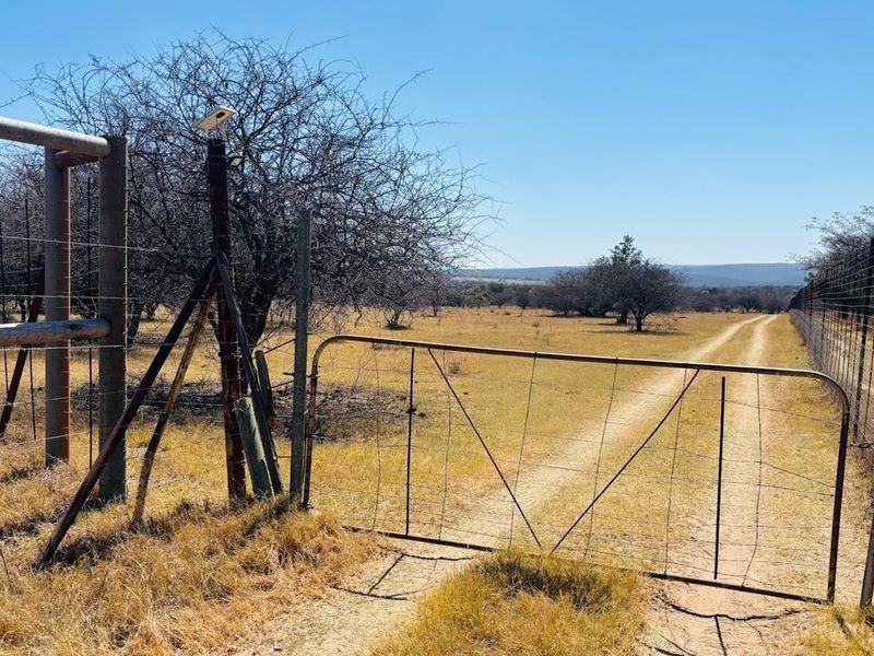 A rural farm entrance featuring a metal gate, wire perimeter fencing, and a dirt access track leading into the open landscape.