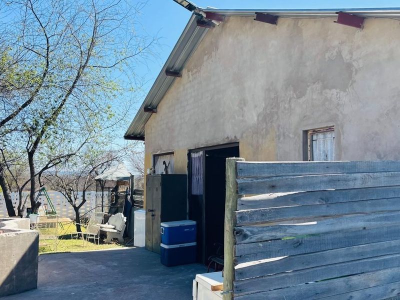 Rear exterior view of a farm building featuring a concrete patio area, metal roofing, and timber fencing, showing signs of significant weathering.