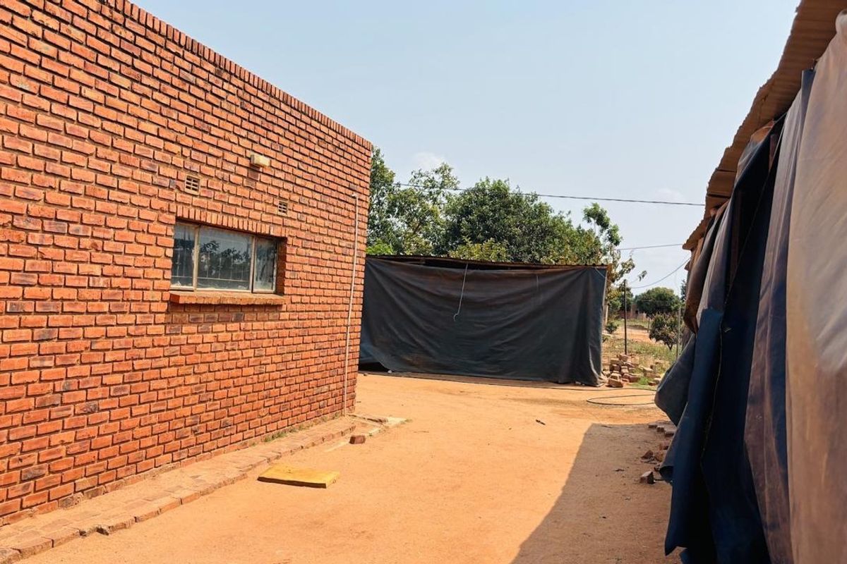 Rear exterior view of a brick house featuring an exposed masonry wall and aluminium window frame, with an unpaved yard area.