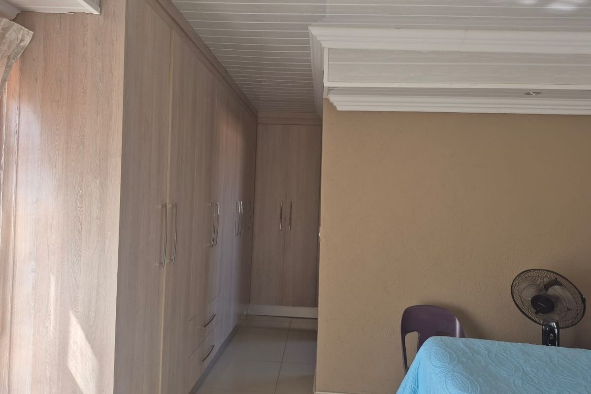 A bedroom featuring extensive built-in cupboards with light wood-grain finish, tiled flooring, and a white tongue and groove ceiling with decorative cornice.