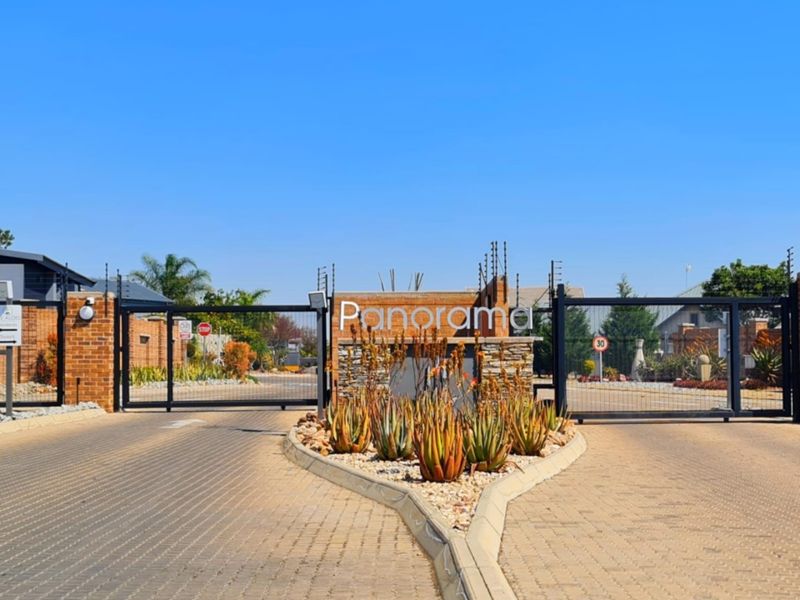 The secure entrance to Panorama estate featuring a paved driveway, electric gates, brick boundary walls, and well-maintained landscaping.