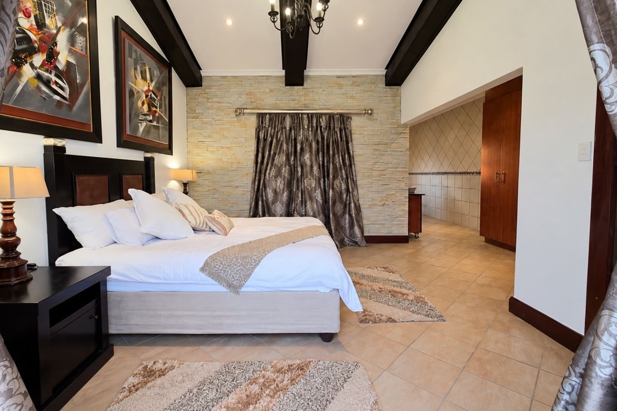 Spacious bedroom featuring exposed timber ceiling beams, a stone feature wall, tiled flooring, and integrated built-in cupboards with an adjoining dressing area.