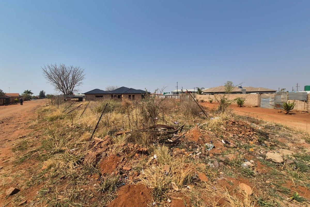 A view of a vacant residential plot featuring unpaved road access and perimeter fencing, situated in a developing residential area.
