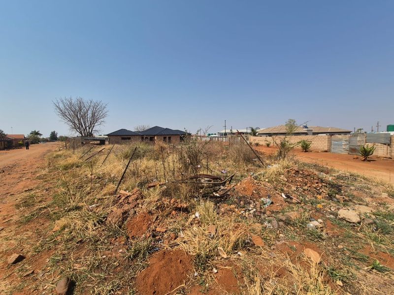 A view of a vacant residential plot featuring unpaved road access and perimeter fencing, situated in a developing residential area.