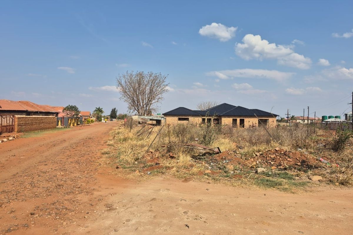 A street-level view of a residential plot featuring an unpaved road, perimeter fencing, and a partially constructed dwelling in a developing neighborhood.