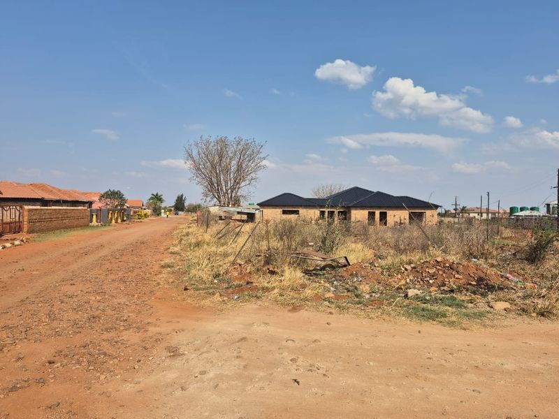 A street-level view of a residential plot featuring an unpaved road, perimeter fencing, and a partially constructed dwelling in a developing neighborhood.