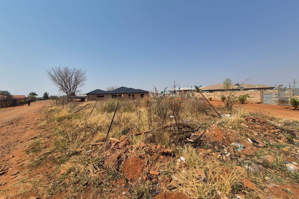 A vacant residential plot featuring dirt road access and wire fencing, currently covered in dry, overgrown vegetation and debris.