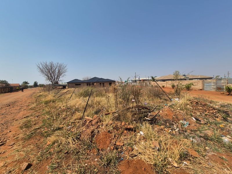 A vacant residential plot featuring dirt road access and wire fencing, currently covered in dry, overgrown vegetation and debris.