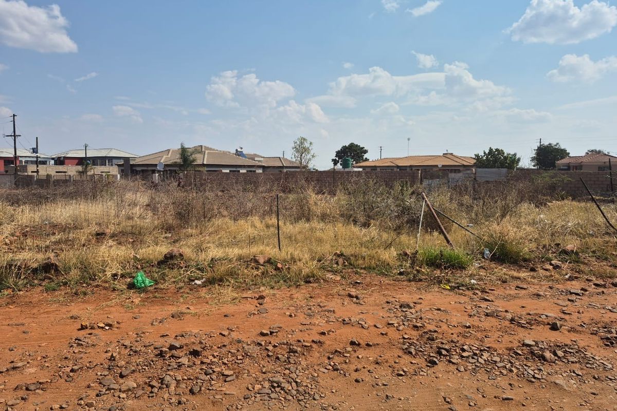 A vacant residential plot featuring dry, overgrown vegetation and perimeter fencing, accessible via an unpaved road in a developed neighborhood.