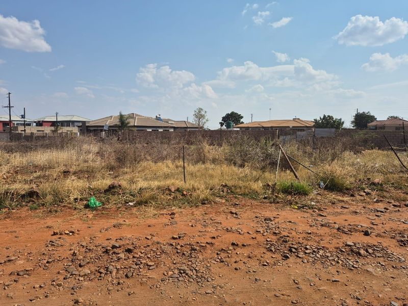 A vacant residential plot featuring dry, overgrown vegetation and perimeter fencing, accessible via an unpaved road in a developed neighborhood.