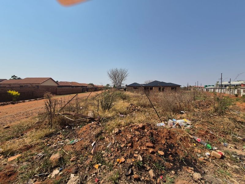 A vacant residential plot featuring natural terrain and dirt road access, situated in an established neighborhood setting.