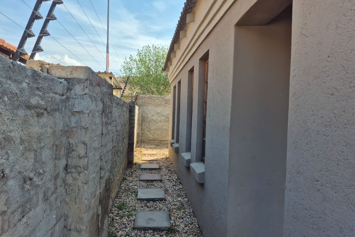 A narrow side passage featuring a gravel path with stone pavers, bordered by a plastered exterior wall and a weathered boundary wall with electric fencing.