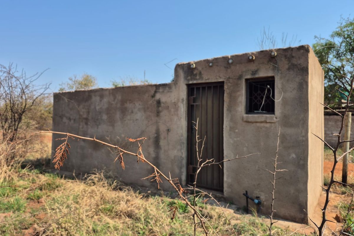 A small, detached utility structure featuring a metal security door and window grille, situated on a vacant residential plot.