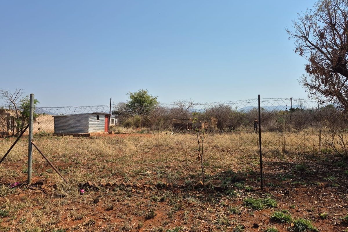 A view of a vacant residential plot featuring dry vegetation, perimeter barbed wire fencing, and a small corrugated metal structure.