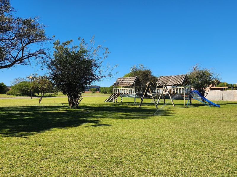 A spacious residential garden featuring a well-maintained lawn, mature trees, and a wooden playground structure with a slide.