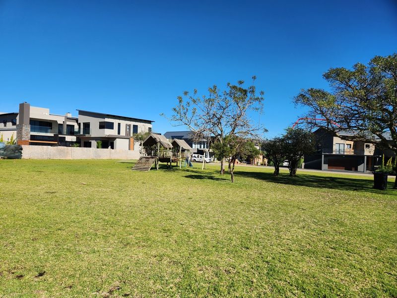 A spacious, well-maintained residential garden featuring a manicured lawn, mature trees, and a secure perimeter wall in a quiet neighborhood setting.