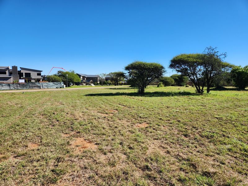 A vacant residential plot featuring open grassland and mature trees, situated in a developed neighborhood setting.