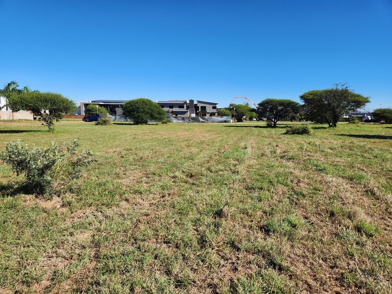 A vacant residential plot featuring open grassland and native trees, situated in a developing neighborhood with surrounding modern residential properties.