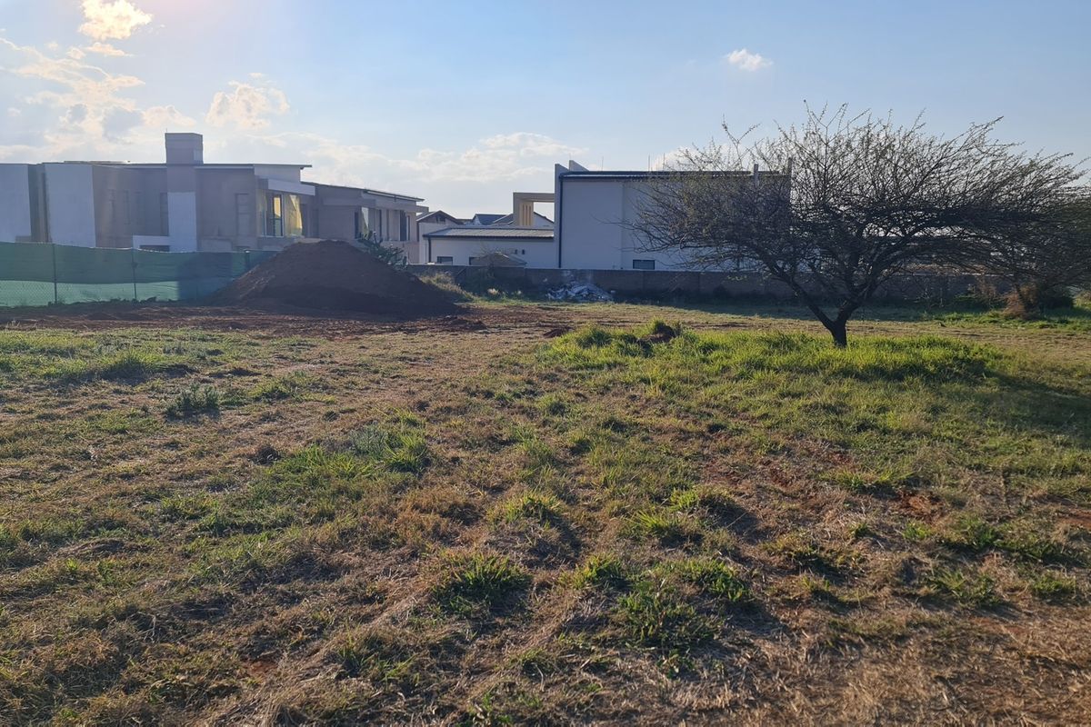 A view of a vacant residential plot featuring natural grass, a mature tree, and surrounding boundary walls in a developed estate.