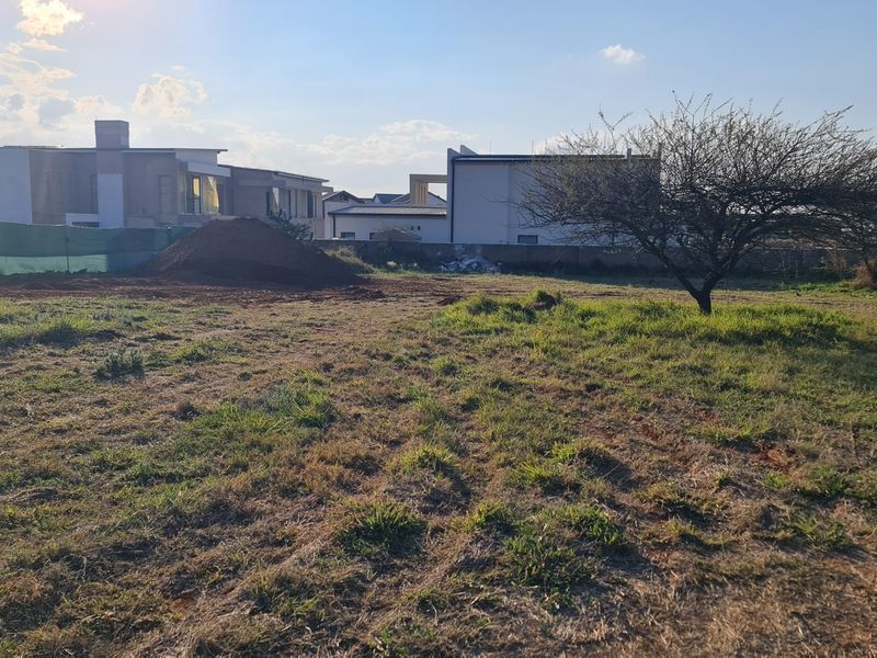 A view of a vacant residential plot featuring natural grass, a mature tree, and surrounding boundary walls in a developed estate.