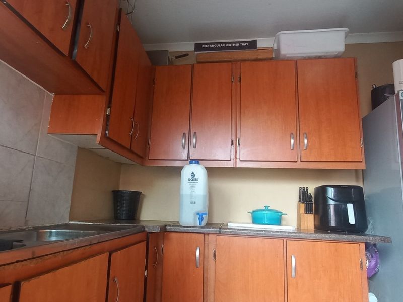 A compact kitchen featuring dated wooden cabinetry, laminate countertops, and a double stainless steel sink with tiled backsplash.