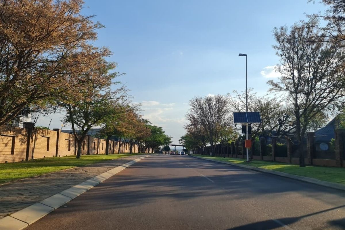 A view of a well-maintained residential street featuring paved roads, brick boundary walls with electric fencing, and solar-powered street lighting.