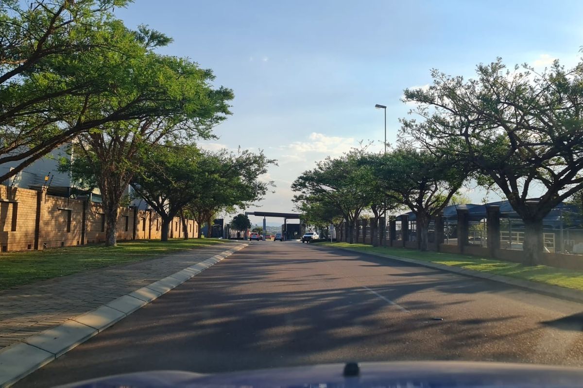 A view of the paved residential street within the estate, featuring mature trees, brick boundary walls, and well-maintained verges leading to the entrance gate.