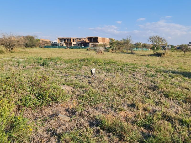 A view of a vacant residential plot of land with natural grass cover and surrounding residential development in the background.