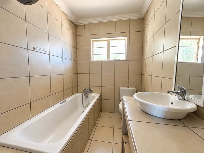 A functional bathroom featuring a built-in bathtub, a vanity basin with a chrome mixer tap, and neutral ceramic tiling throughout.